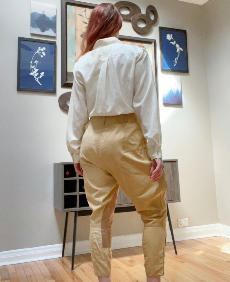 May include: A cream-colored long-sleeved button-down shirt paired with tan riding pants. The pants have a high waist, a fitted lower leg, and a suede-like patch on the outer thigh. The outfit is displayed indoors.