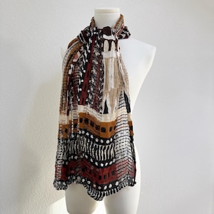 Può includere: Sciarpa fantasia drappeggiata su un manichino, nei toni del marrone, nero e bianco. La sciarpa presenta un design geometrico con rettangoli, punti e linee. Realizzata in tessuto leggero.