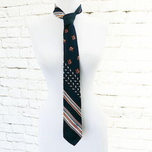 May include: A dark navy tie with a repeating paisley pattern and diagonal stripes in orange, white, and blue. The tie is displayed on a white mannequin against a white brick wall.