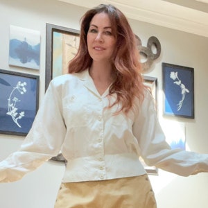 May include: Cream-colored button-down shirt with long sleeves. The shirt has a classic collar and a fitted waist. The fabric appears to have a subtle texture. The model is wearing a tan skirt.