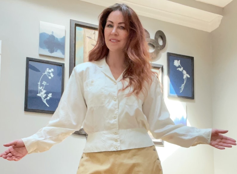 May include: Cream-colored button-down shirt with long sleeves. The shirt has a classic collar and a fitted waist. The fabric appears to have a subtle texture. The model is wearing a tan skirt.