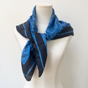 May include: A square scarf featuring a vibrant blue center with a black cursive script pattern, bordered by a wide black band and a thin blue stripe. The scarf is tied in a knot at the front, displaying a smooth fabric texture and a bold color contrast.