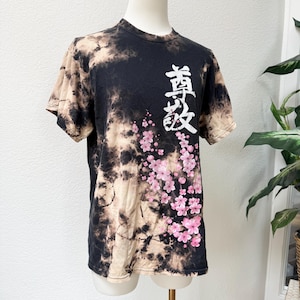 May include: Black and tan tie-dye t-shirt with white Japanese characters and pink cherry blossoms. The short-sleeved shirt is displayed on a mannequin. The design features a floral pattern and text.