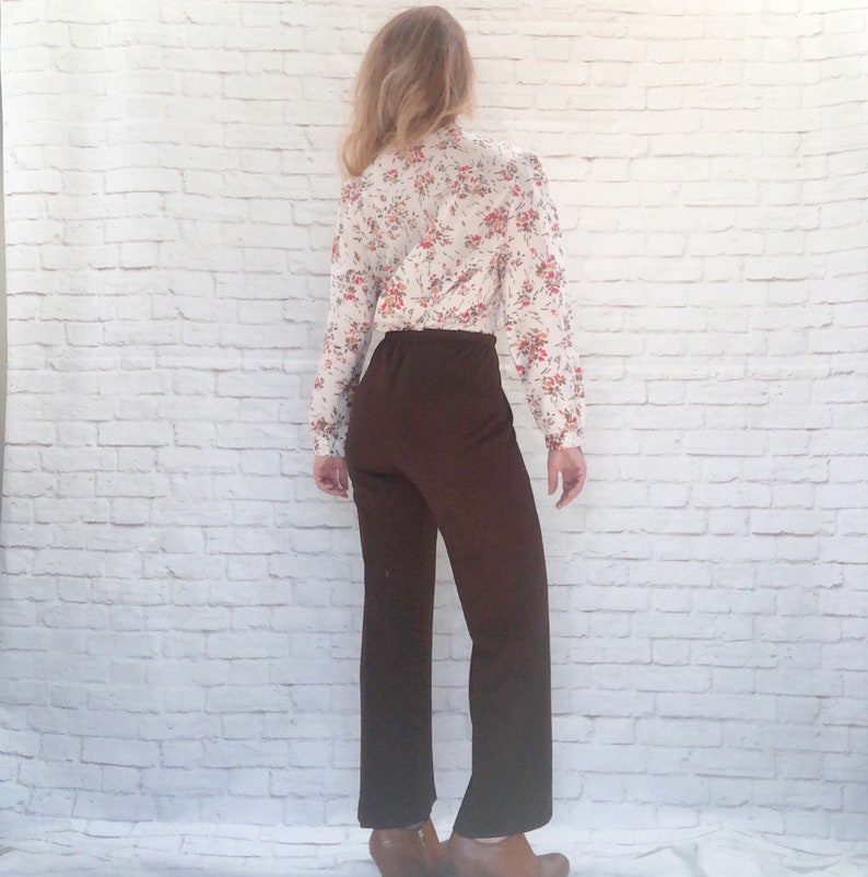 May include: A vintage outfit featuring a floral print blouse and brown pants. The long-sleeved blouse has a white background with red and orange floral patterns. The high-waisted pants are a solid dark brown color.