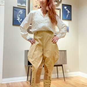 May include: A woman wearing a white collared shirt and tan breeches. The breeches have a high waist and are gathered at the knees, with button details. The outfit is styled in a vintage or historical fashion.