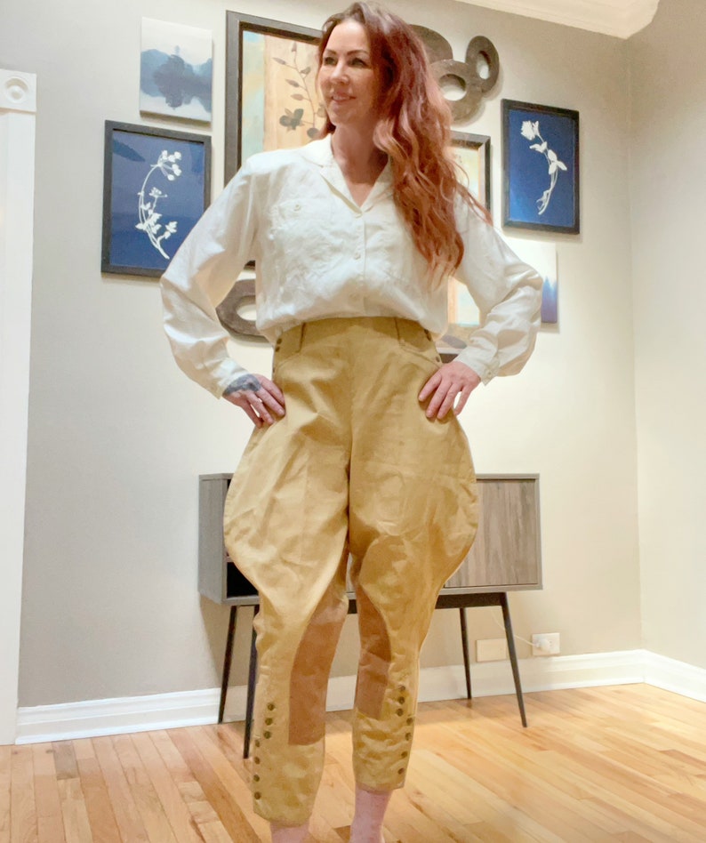 May include: A woman wearing a white collared shirt and tan breeches. The breeches have a high waist and are gathered at the knees, with button details. The outfit is styled in a vintage or historical fashion.