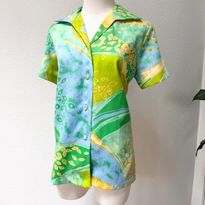 May include: A short-sleeved button-down shirt featuring a vibrant patchwork pattern in shades of lime green, sky blue, and yellow. The design includes abstract leaf motifs, watercolor-style washes, and crackled textures across the fabric.