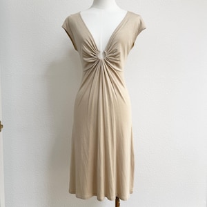 May include: A beige cap-sleeved dress with a V-neck and gathered detailing at the bust. The dress is displayed on a mannequin and has a knee-length hemline. The fabric appears to be a soft, flowing material.
