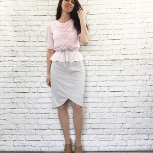 May include: A light pink blouse with short sleeves and a cowl neckline, paired with a gray pencil skirt. The outfit is cinched at the waist with a gray belt. The model is wearing brown platform sandals.