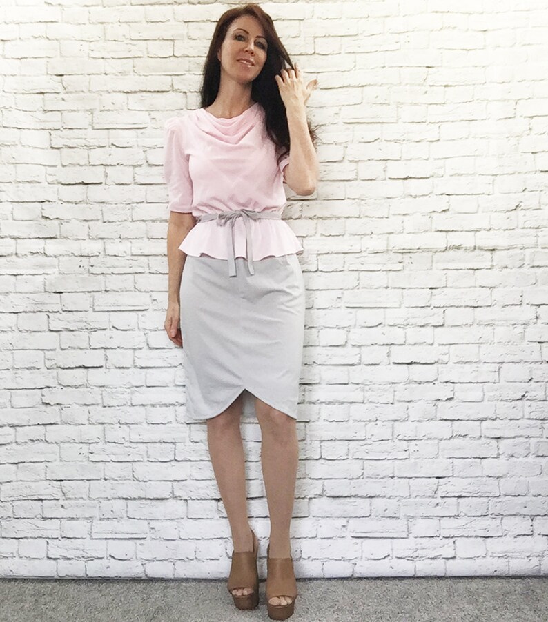 May include: A light pink blouse with short sleeves and a cowl neckline, paired with a gray pencil skirt. The outfit is cinched at the waist with a gray belt. The model is wearing brown platform sandals.