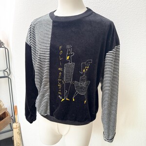 May include: Black long-sleeve sweatshirt with a striped panel on the left side and sleeves. The sweatshirt features an embroidered design of two figures and the text "FOLIE ELECTRIQUE" in yellow.
