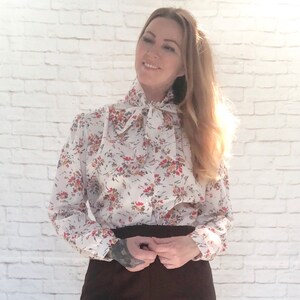 May include: A white blouse with a floral pattern in red, orange, and brown. The blouse has a high, ruffled collar and a tie at the neck. The sleeves are long and gathered at the cuffs. The blouse is paired with a dark brown skirt.