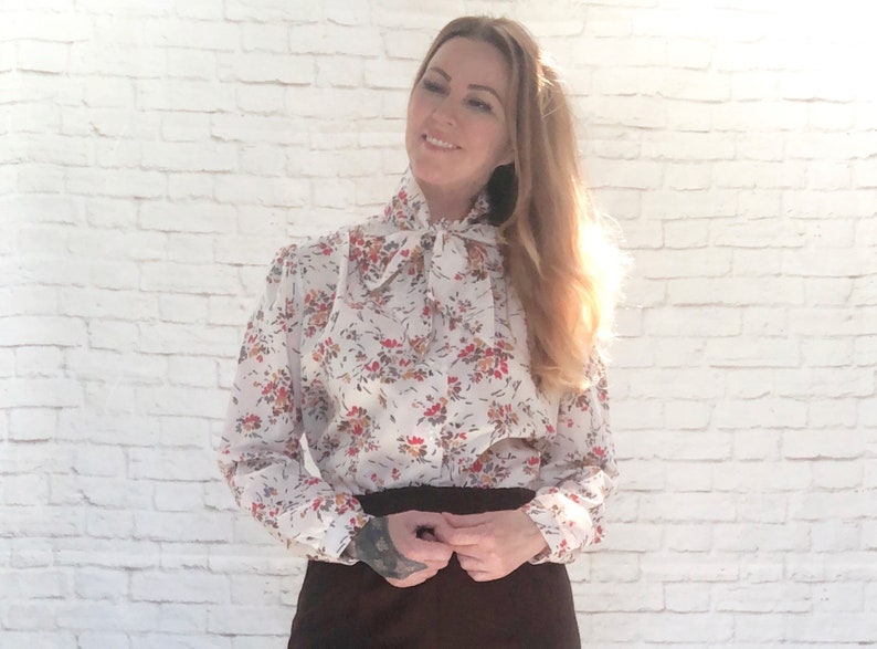 May include: A white blouse with a floral pattern in red, orange, and brown. The blouse has a high, ruffled collar and a tie at the neck. The sleeves are long and gathered at the cuffs. The blouse is paired with a dark brown skirt.