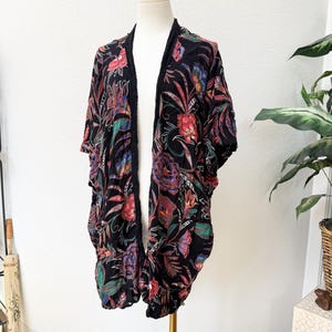 Może przedstawiać: Czarny kimono z żywym kwiatowym wzorem w kolorze czerwonym, niebieskim i zielonym. Otwarta odzież ma czarne wykończenie. Tkanina wydaje się lekka i zwiewna, idealna do warstw. Prezentowane na manekinie.
