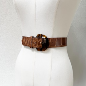 May include: A brown belt with a faux crocodile texture. The belt features a large, oval-shaped buckle with a tortoiseshell pattern. The belt is displayed on a white mannequin.