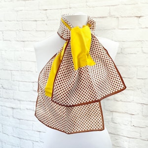May include: A yellow and brown scarf with a geometric pattern is draped over a white mannequin. The scarf has a diamond pattern in brown and white, with a solid yellow section. The edges are trimmed in brown.