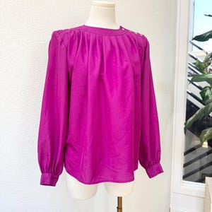 May include: A vibrant magenta blouse with long sleeves and a round neckline. The blouse features shoulder pleats and decorative buttons on the shoulder. The sleeves have buttoned cuffs. The blouse is displayed on a mannequin.