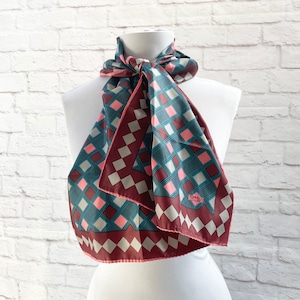 May include: A patterned scarf in shades of teal, burgundy, pink, and gray, displayed on a white mannequin. The scarf features a geometric diamond design and a border. The scarf is knotted at the neck, showcasing its length and pattern.