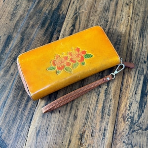 May include: A rectangular mustard yellow wallet featuring an embossed floral design of two orange hibiscus flowers with green leaves. The wallet includes a silver-toned metal zipper closure and a detachable brown leather wrist strap with a metal clip.