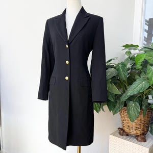 Puede incluir: Un vestido blazer negro con cuello con solapas y mangas largas. El vestido tiene tres botones dorados y dos bolsillos delanteros. El vestido se exhibe en un maniquí.