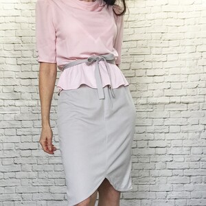 May include: A light pink blouse with short sleeves and a round neckline, paired with a gray skirt. The blouse has a peplum detail and a gray tie belt. The skirt is knee-length and has a tulip-style hem.