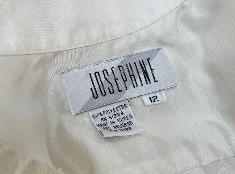 May include: Close-up of a white label with the name "JOSEPHINE" in black capital letters. The label also includes the size "12" and text indicating the material is 100% polyester and made in Korea.
