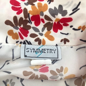 May include: Close-up of a cream-colored fabric with a floral pattern in red, brown, gray, and yellow. The fabric has a label that reads "SYMMETRY". The floral design includes black stems and leaves.