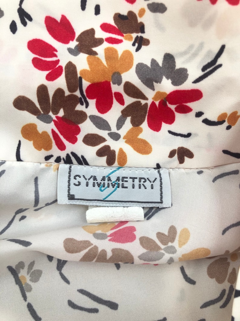 May include: Close-up of a cream-colored fabric with a floral pattern in red, brown, gray, and yellow. The fabric has a label that reads "SYMMETRY". The floral design includes black stems and leaves.