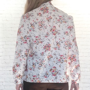 May include: A white blouse with a floral pattern in red, brown, and yellow. The long-sleeved shirt has a high collar and buttoned cuffs. The floral print is scattered across the fabric.