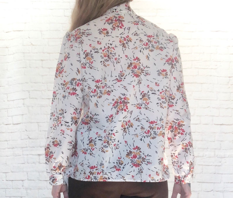 May include: A white blouse with a floral pattern in red, brown, and yellow. The long-sleeved shirt has a high collar and buttoned cuffs. The floral print is scattered across the fabric.