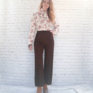 May include: A vintage outfit featuring a cream-colored blouse with a floral pattern in red and orange, and a brown pair of wide-leg pants. The blouse has a bow at the neck. The pants are high-waisted and flared. The outfit is paired with platform heels.