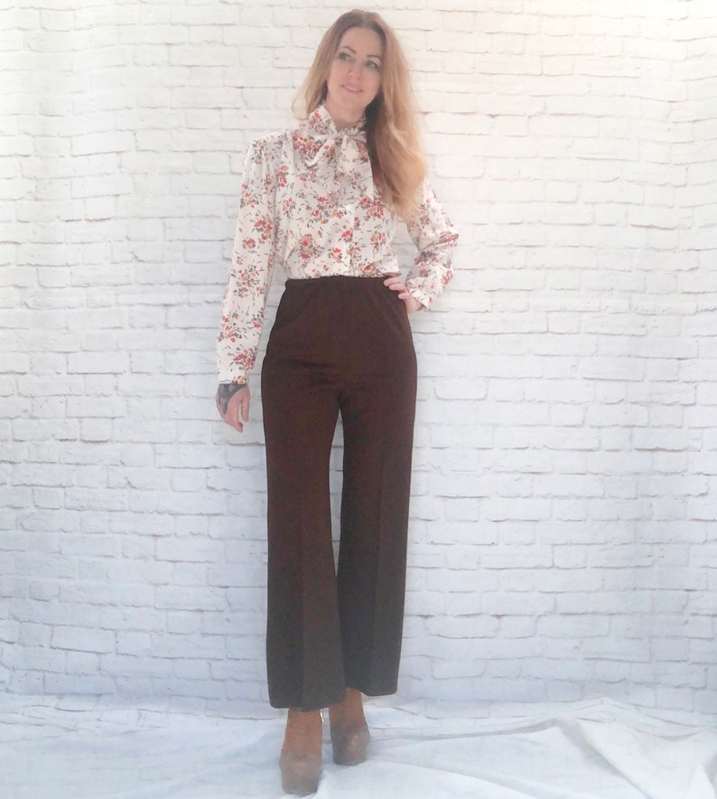 May include: A vintage outfit featuring a cream-colored blouse with a floral pattern in red and orange, and a brown pair of wide-leg pants. The blouse has a bow at the neck. The pants are high-waisted and flared. The outfit is paired with platform heels.