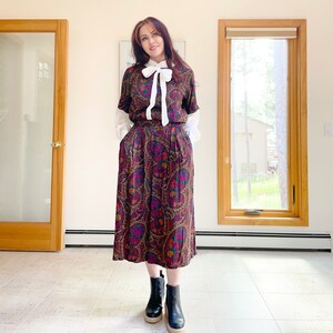May include: A vintage paisley print dress with a white bow and sleeves. The dress features a midi skirt and is paired with black platform boots. The paisley pattern includes shades of purple, gold, and green.