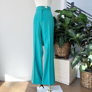 Può includere: Pantaloni a vita alta color teal con gamba ampia. I pantaloni sono esposti su un manichino, mostrando la vestibilità su misura e la chiusura con bottoni in vita. I pantaloni sono realizzati in un tessuto liscio.