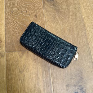 May include: A rectangular black wallet featuring a glossy, embossed crocodile skin texture. The wallet is secured with a wrap-around zipper that includes a small, rectangular gold-toned metal pull tab attached to the side of the accessory.
