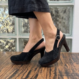 Op de afbeelding: Een paar zwarte suède slingback-hakken met een ronde neus, een geweven textuurdetail aan de zijkant, een donkerbruine houten plateauzool en een blokhak met een verstelbare zilverkleurige metalen gespsluiting.