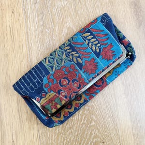 May include: A rectangular clutch with a metal clasp. The fabric is a patchwork of blue, teal, red, and yellow floral and geometric patterns. The clutch is closed and lies on a light wood surface.