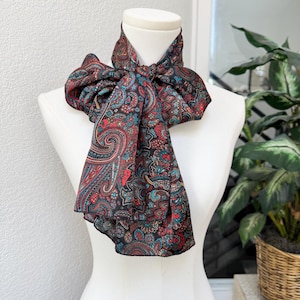 May include: A paisley patterned scarf in shades of red, teal, and black is draped on a white mannequin. The scarf is tied in a bow at the neck, with the ends cascading down. The intricate design features swirling patterns and floral motifs, creating a luxurious accessory.