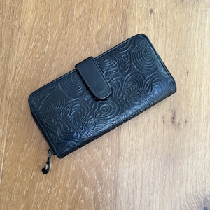 May include: A black rectangular wallet featuring an embossed swirling pattern across its surface. The wallet includes a secure snap-button closure flap on the front and a metal zipper pull attached to the side, providing a functional design for storage.