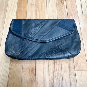 Op de afbeelding: Een zwarte leren clutch met een patchwork ontwerp. De clutch heeft een klepsluiting en is gemaakt van verschillende texturen zwart leer, waaronder gladde en getextureerde panelen. De stiksels zijn zichtbaar en dragen bij aan het ontwerp.