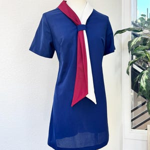 May include: A short-sleeved navy blue dress featuring a V-neckline with a decorative tie detail. The tie consists of two long, flat fabric strips in solid white and deep red, secured at the center of the neckline, hanging down the front.