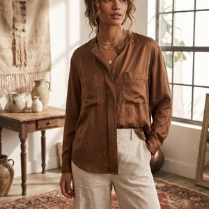 May include: A long-sleeved, button-down shirt in a solid brown satin-like fabric featuring a mandarin collar, two front chest pockets, and a relaxed fit, paired with high-waisted, wide-leg trousers in a light beige, textured linen-like material.