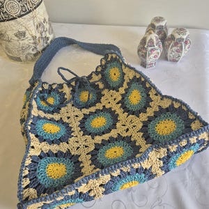 May include: A handmade crochet tote bag in a floral pattern with blue, yellow, and cream colors. The bag has a blue strap and a drawstring closure. Decorative ceramic jars with floral patterns are in the background.