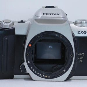 May include: A silver and black Pentax ZX-50 camera. The camera has a lens mount, a viewfinder, and various control dials. The Pentax logo is visible on the top and side of the camera. The camera is designed for photography.