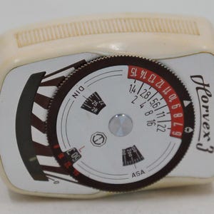 May include: A vintage ivory-colored Horvex 3 light meter. The meter features a circular dial with black and red markings for settings like DIN and ASA. The name "Horvex 3" is printed in black on the side.
