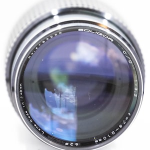 May include: A close-up of a Soligor camera lens with the text "SOLIGOR" and "Made in Japan". The lens has a silver metal body and a glass lens with a blue reflection. The lens has the following specifications: f=78~210mm, 1:3.5, and 62mm.