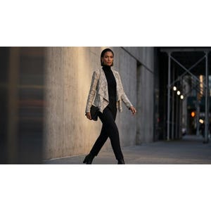 Puede incluir: Un blazer con estampado de piel de serpiente en tonos beige y gris con frente abierto, usado sobre un suéter de cuello alto negro y pantalones negros. El conjunto incluye un bolso de mano negro y botines negros, creando un estilo coordinado.