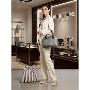 Puede incluir: Un bolso gris estructurado con textura tejida y asas de cuero negro, sostenido por una persona que viste un blazer y pantalones color crema. Una bolsa de tela a rayas marrones y blancas se encuentra en el suelo en primer plano.