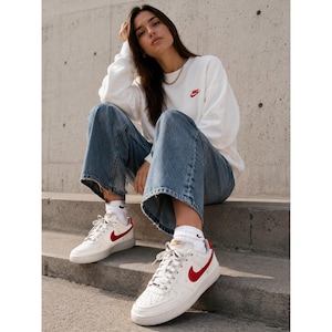 Puede incluir: Zapatillas Nike Air Force 1 blancas con detalles rojos, combinadas con vaqueros azules y una sudadera blanca con un pequeño logo Nike rojo. Las zapatillas tienen un diseño clásico de corte bajo con un swoosh rojo.