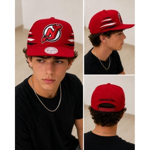 Puede incluir: Una gorra de béisbol roja con el logotipo de los New Jersey Devils bordado en el frente en rojo, negro y blanco. Los lados muestran gráficos de marcas de garras en negro y blanco. La parte posterior incluye un cierre ajustable y texto Mitchell & Ness.